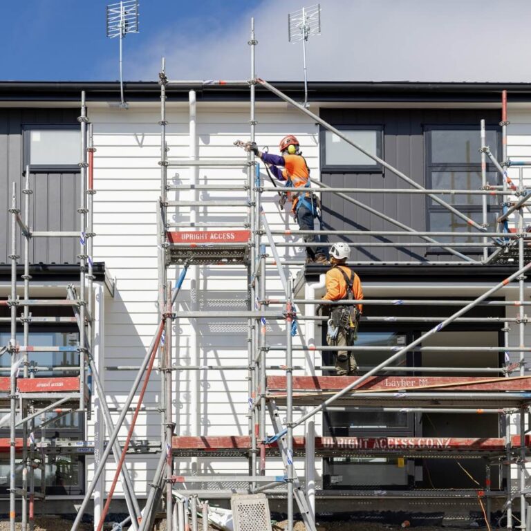 Two scaffolding apprentices in hi-vis and helmets assembling scaffolding on a building site.
