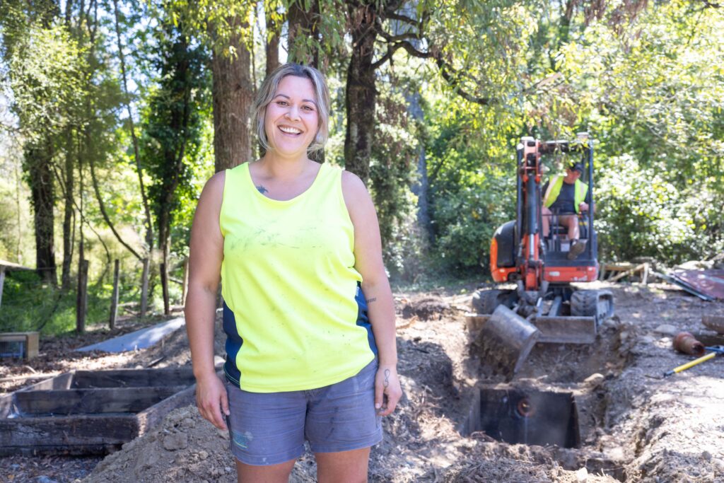 Apprentice in hi-vis singlet smiling on site with excavation machinery in the background